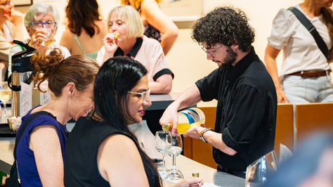 A busy wine event shows a man with curly hair pouring white wine into glasses. Attendees chat and enjoy drinks, creating a lively and social atmosphere.