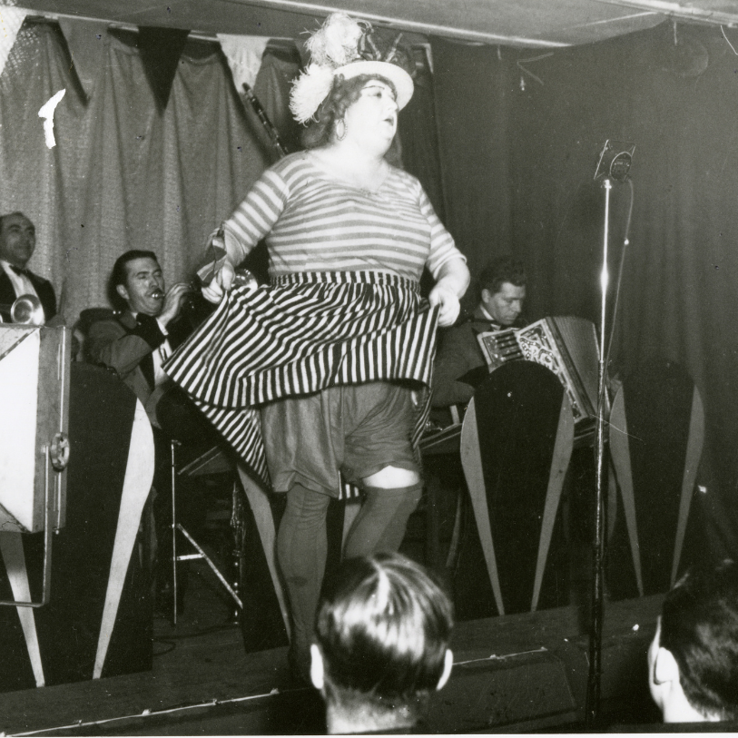 A drag performer on stage with a band playing instruments behind them.