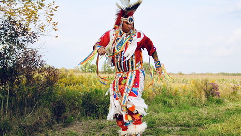 An Indigenous dancer in vibrant traditional regalia performs outdoors in a grassy field during a cultural dance demonstration.