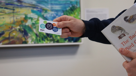 A person holds an HHPL library card and a bird identification book in front of a colourful painting inside the library.
