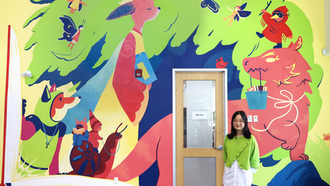 Artist April Zheng stands in front of a colourful mural inside the library featuring whimsical woodland animals, fairies, birds, and books beneath a large green tree.