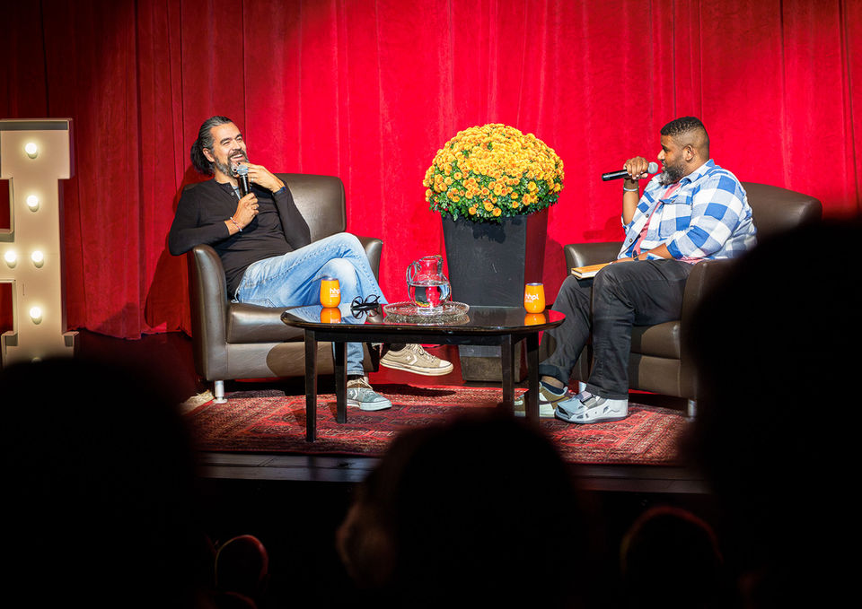 Two people speaking on a stage with red curtains.