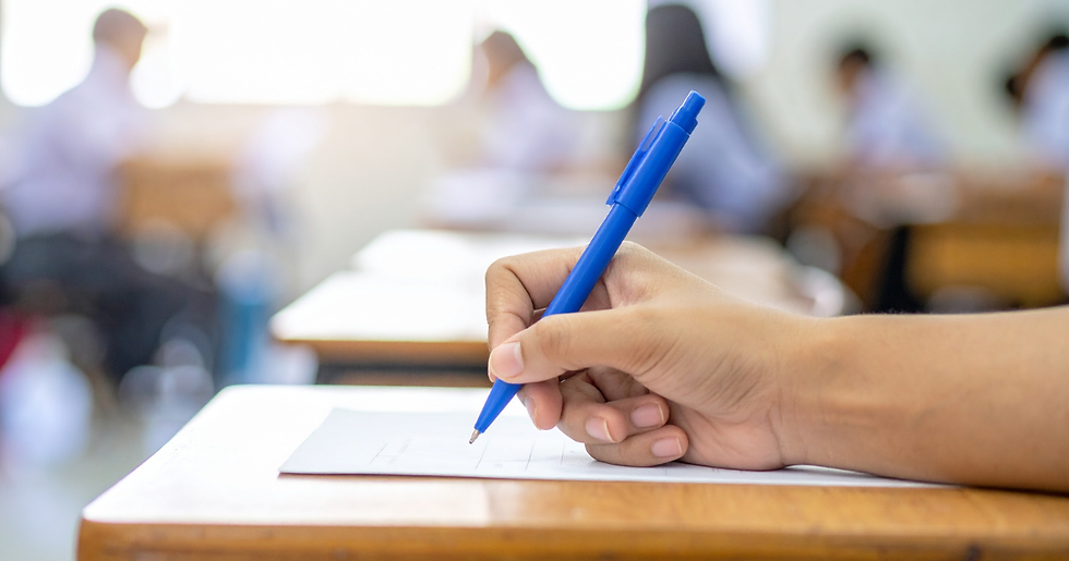 A person writing on a test paper in a classroom with other students blurred in the background.
